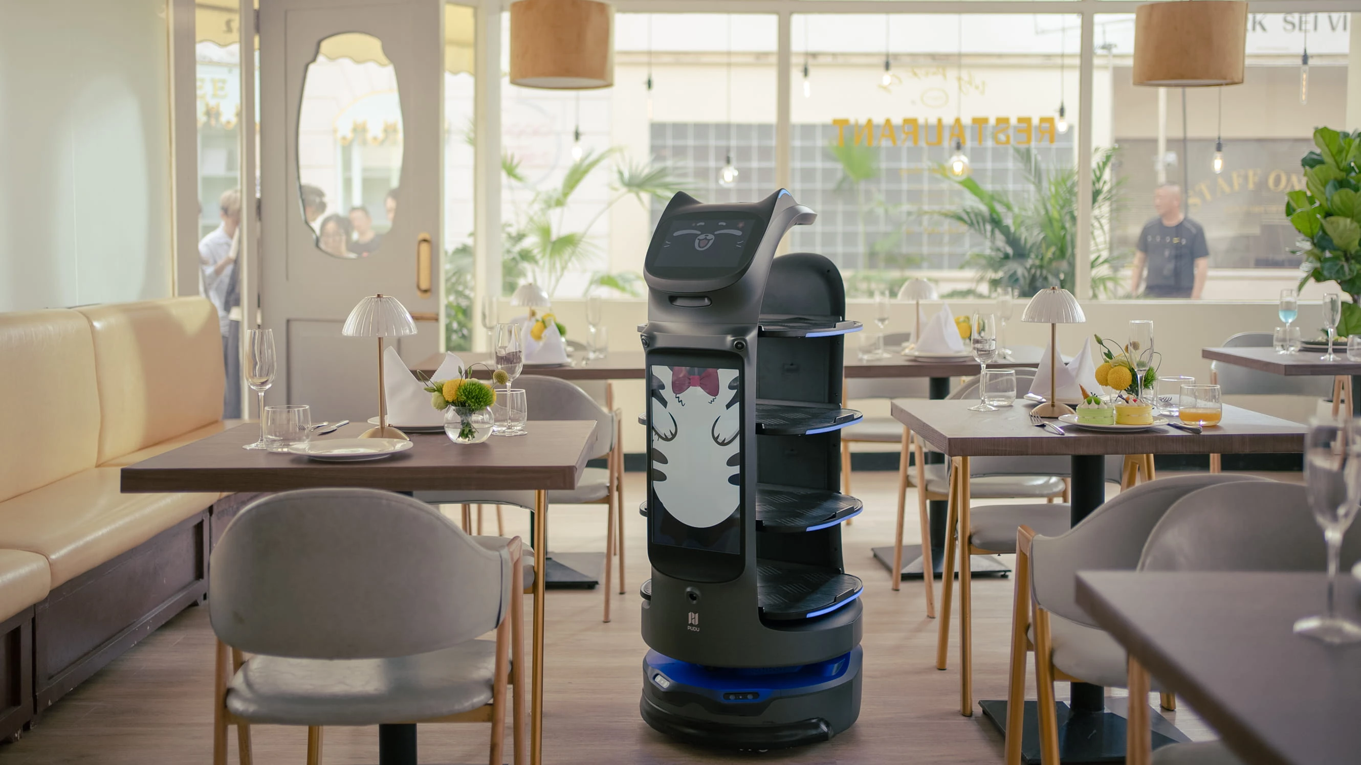 Restoran salonunda masaların arasında duran kedi yüzlü servis robotu